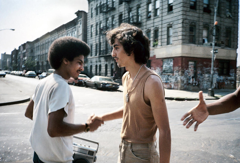 Meryl Meisler. Handshake. 1984. Image courtesy of the artist. Alt text: Two young men with mustaches shake each others hand in greeting on a New York City street. The man on the left wears a white t-shirt and holds a silver boombox. The man on the right wears a beige tank top tucked into slim khaki pants with a silver chain necklace around his neck. On the right side of the photograph, an outstretched hand reaches towards them from outside of the frame.