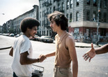 Meryl Meisler. Handshake. 1984. Image courtesy of the artist. Alt text: Two young men with mustaches shake each others hand in greeting on a New York City street. The man on the left wears a white t-shirt and holds a silver boombox. The man on the right wears a beige tank top tucked into slim khaki pants with a silver chain necklace around his neck. On the right side of the photograph, an outstretched hand reaches towards them from outside of the frame.
