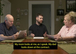 Shared Resources. 2021. Directed by Jordan Lord. Courtesy the filmmaker. Image description: Three white people sit at a table together, each with a set of white papers in front of them. The figure on the far left is a bald man in his 60s, who reads the papers intently. The figure in the center is a person in their late 20s, who looks to a woman in her 60s with blonde hair, who holds the papers in her hands and looks back at them. A caption appears over the image that reads: “My mom looks at me, as I speak. My dad looks down at the contract.”
