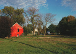 Amanda Williams. Harold&#39;s Chicken Shack, from the Color(ed) Theory Suite. 2014–2016. Pigmented inkjet print. Fund for the Twenty-First Century Alt Text: A photograph of a large grassy yard under a cloud-dotted blue sky. A concrete path winds across the yard, ending on the left at a brightly colored but worn orange-red house with no windows and a fence covered in overgrowth. Centered in the background is another wooden house, hidden behind trees and surrounded by a few parked cars. Large green and brown leafy hedges frame this residential landscape on the left and right sides, and the long shadows of trees streak across the yard.