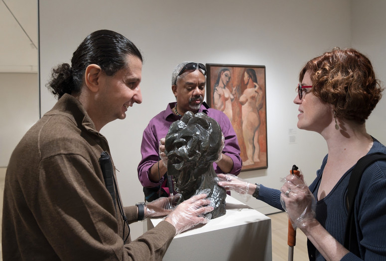 A horizontally oriented photograph of three museum visitors touching a bumpy bronze bust by artist Pablo Picasso on a white pedestal in a gallery. They wear plastic gloves and discuss the sculpture. Photo: Martin Seck. ©️ 2023 The Museum of Modern Art, New York