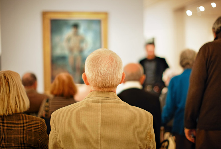 Photo: Jason Brownrigg ©️ 2024 The Museum of Modern Art, NY. Image description: A man in a tan corduroy jacket is centered among a blurred group of adults sitting with their backs turned to look at a framed artwork on the wall.