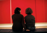 Photo: Jason Brownrigg ©️ 2024 The Museum of Modern Art, NY. Image description: Two women sit with their backs turned to the viewer, looking at a red canvas with vertical lines that hangs the whole span of the wall.