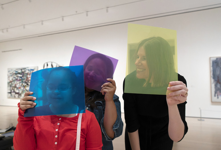Photo: Martin Seck. © 2024 The Museum of Modern Art, New York Image description: A smiling girl in a red shirt holds a sheer blue square in front of her face alongside two smiling women holding purple and yellow squares.