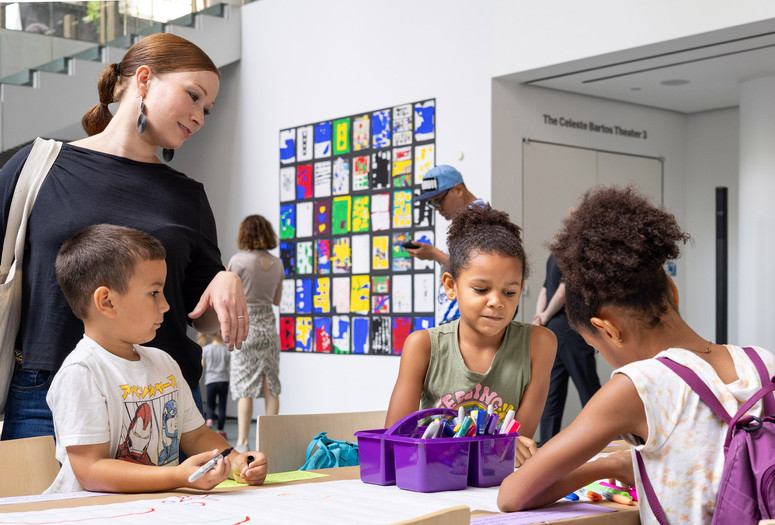 Photo: onwhitewall.com. © 2024 The Museum of Modern Art, NY Image Description: Three children draw at a table covered with coloring paper and a purple box of markers while a red-haired woman in a black dress looks over them and smiles.