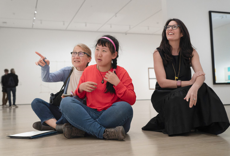 Photo: Martin Seck. © 2023 The Museum of Modern Art, New York Image description: A young woman wearing a coral pink shirt and pink headband sits cross-legged on a gallery floor between her mother, pointing off to the left, and a crouched educator in a long black dress looking up in the same direction.
