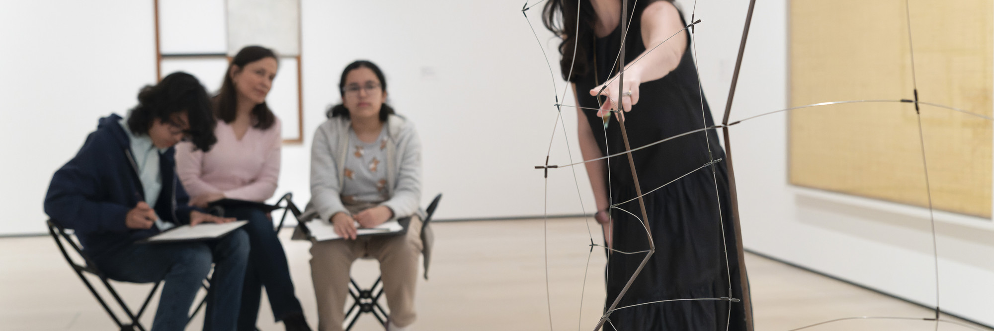 Photo: Martin Seck. © 2023 The Museum of Modern Art, NY Image Description: An educator in a black dress points to a hanging wire sculpture by the artist Gego, while a family of three people sitting on gallery stools look at the artwork and draw together.
