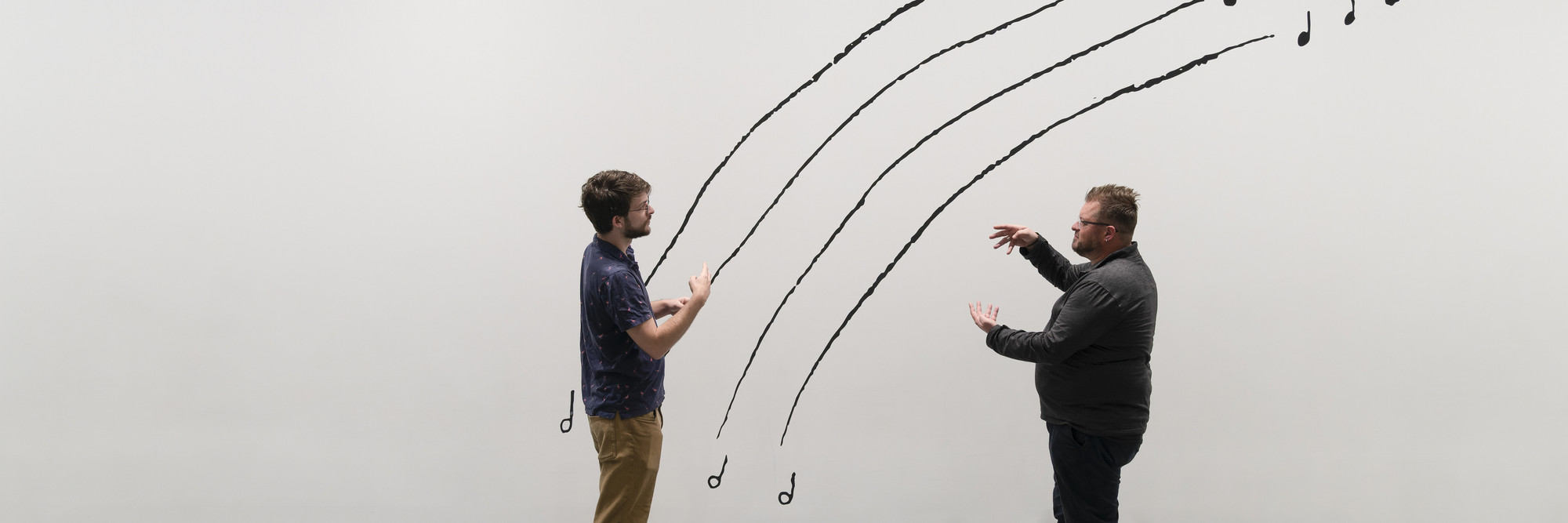 Photo: Martin Seck Image description: Two men sign to each other while standing in front of a black-and-white line drawing mural by the artist Christine Sun Kim. &#34;The Sound of Temperature Rising&#34; is written in capital letters at the top left corner, while the men stand in front of four arching lines that mimic music stanzas.