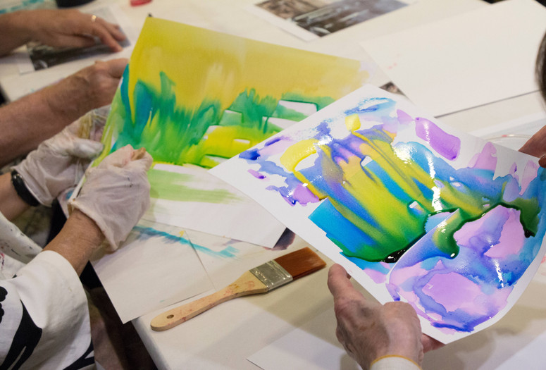 Photo: Manuel Molina Martagon. © 2024 The Museum of Modern Art, New York Image description: A photograph showing the hands of older adults holding watercolor paintings. On the right, the hands hold a wet composition of melding purple, blue, and yellow. To the left, gloved hands paint broad yellow strokes. A wet brush sits on the table between them.