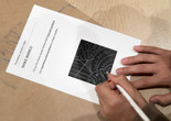 Two hands draw on a small activity sheet, making marks with a white pencil onto a black square background. Photo: Martin Seck