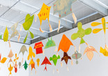 Installation view of Joan Jonas: Good Night Good Morning, The Museum of Modern Art, New York, March 17–July 6, 2024. Photo: Jonathan Dorado. Digital image © 2024 The Museum of Modern Art, New York Image description: An installation of 30 colorful paper kites hanging in three rows from the rafters of a white industrial ceiling. The shapes of the kites are different amorphous forms. They are hand-painted in brilliant yellows, greens, oranges, and reds, with a few brushstrokes in contrasting colors dotting each surface. Some have tufts of tan raffia drifting down from their corners.