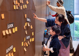 Photo: On White WallImage description: A caregiver points and shows a child written texts on colorful Post-It notes placed on a brown wall.