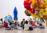 Photo: On White WallImage description: Children in a tour group raise their hands while sitting underneath floating sculptures.