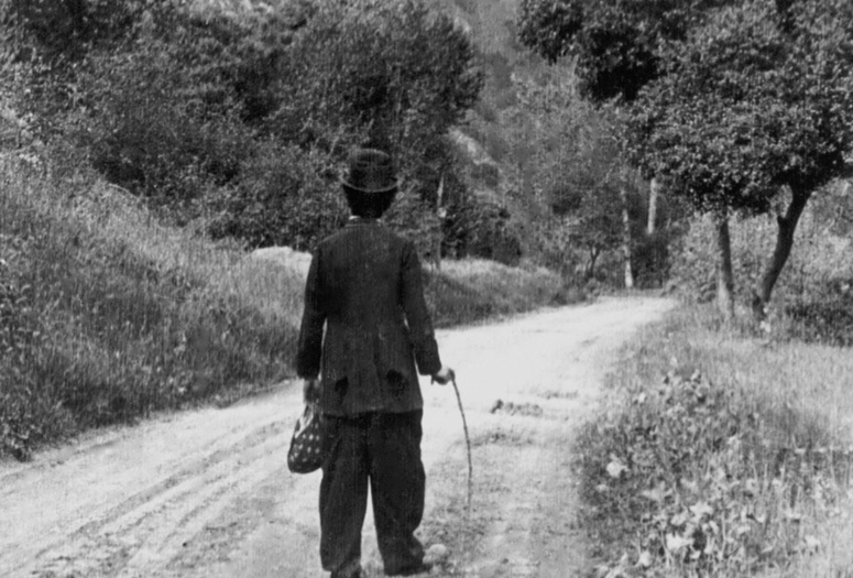 The Tramp. 1915. USA. Written and directed by Charles ChaplinImage description: Black and white film still of a person, with their back to the viewer, wearing a baggy suit and rounded top hat walking up an unpaved road through a woodsy landscape. They hold a long stick as a walking cane.