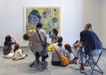 An educator looks up at an artwork depicting houses, and uses her hands as binoculars while she leads a group of families in a conversation. Photo courtesy On White Wall, Inc.