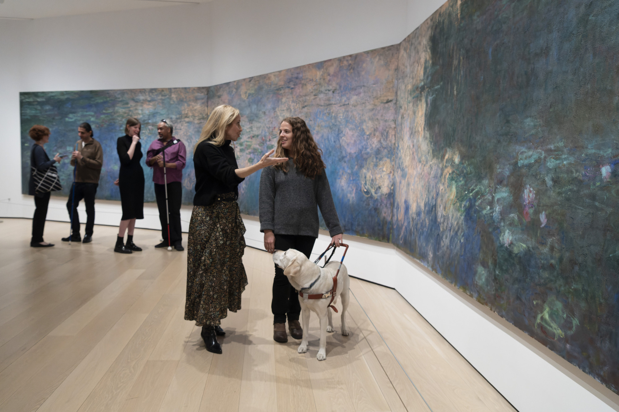 Photo: Martin Seck. ©️ 2023 The Museum of Modern Art, New York  Image description: A group of visitors stand in front of Claude Monet’s painting Water Lilies. The three large connected canvases take up almost the entire wall, with swaths of deep blues, purples, greens, and light pink paint applied in looping strokes across the surface. In the foreground, two women are speaking to each other. One is gesturing in conversation, the other is smiling with her guide dog, who looks up at her. In the background, four people discuss the painting in pairs. Two of them use white canes.