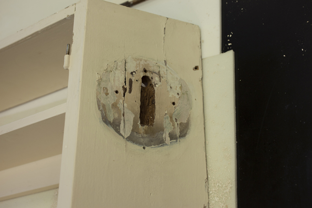 The original small spot light lamp that was installed on the end of this cabinet was replaced at some point with a larger light with a circular base. The side of the cabinet had to be carved back in order accommodate the wiring of the replacement lamp. This image shows the damage to the side of the cabinet. As part of the treatment this damage will be repaired and an example of the original lamp will be installed here.