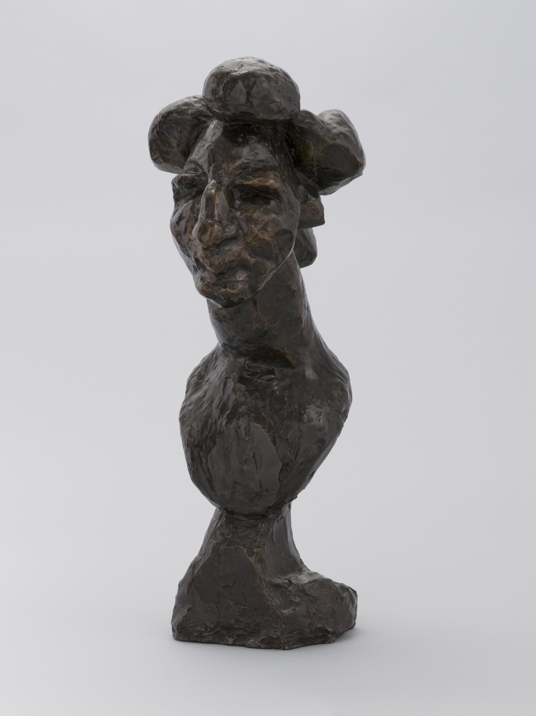 A bronze sculpture with a dark brown, uneven surface depicts a figure's head and shoulders, featuring exaggerated facial features. The background is plain and light grey. This is an AI generated description. To give feedback, email ai@moma.org.
