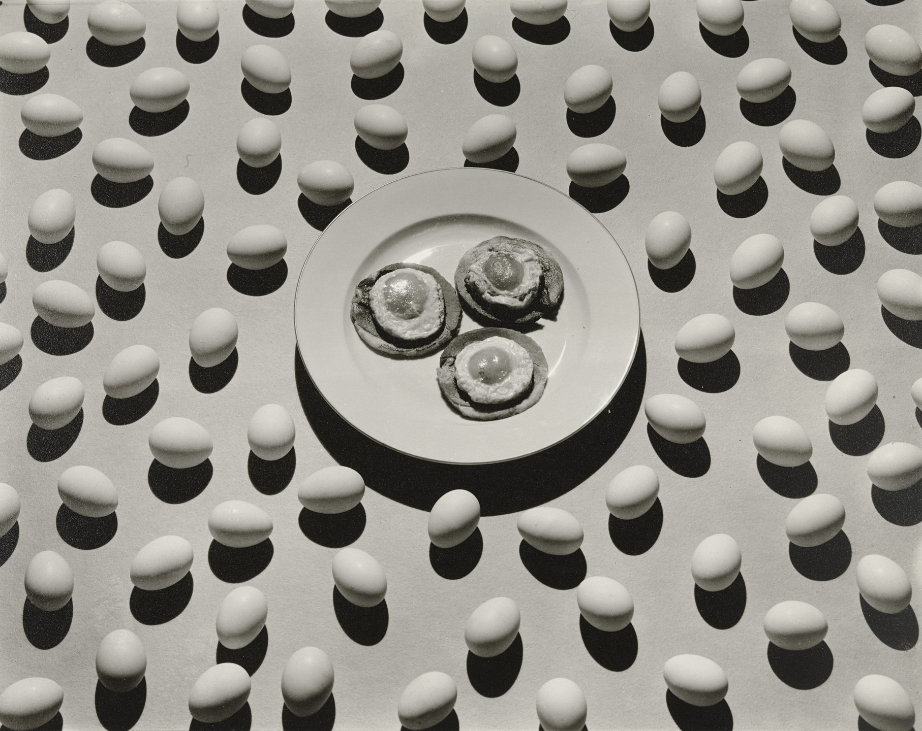 A white plate with three pastries or buns in the center is surrounded by a pattern of evenly spaced white eggs, casting shadows on a light background. The image is in black and white. This is an AI generated description. To give feedback, email ai@moma.org.