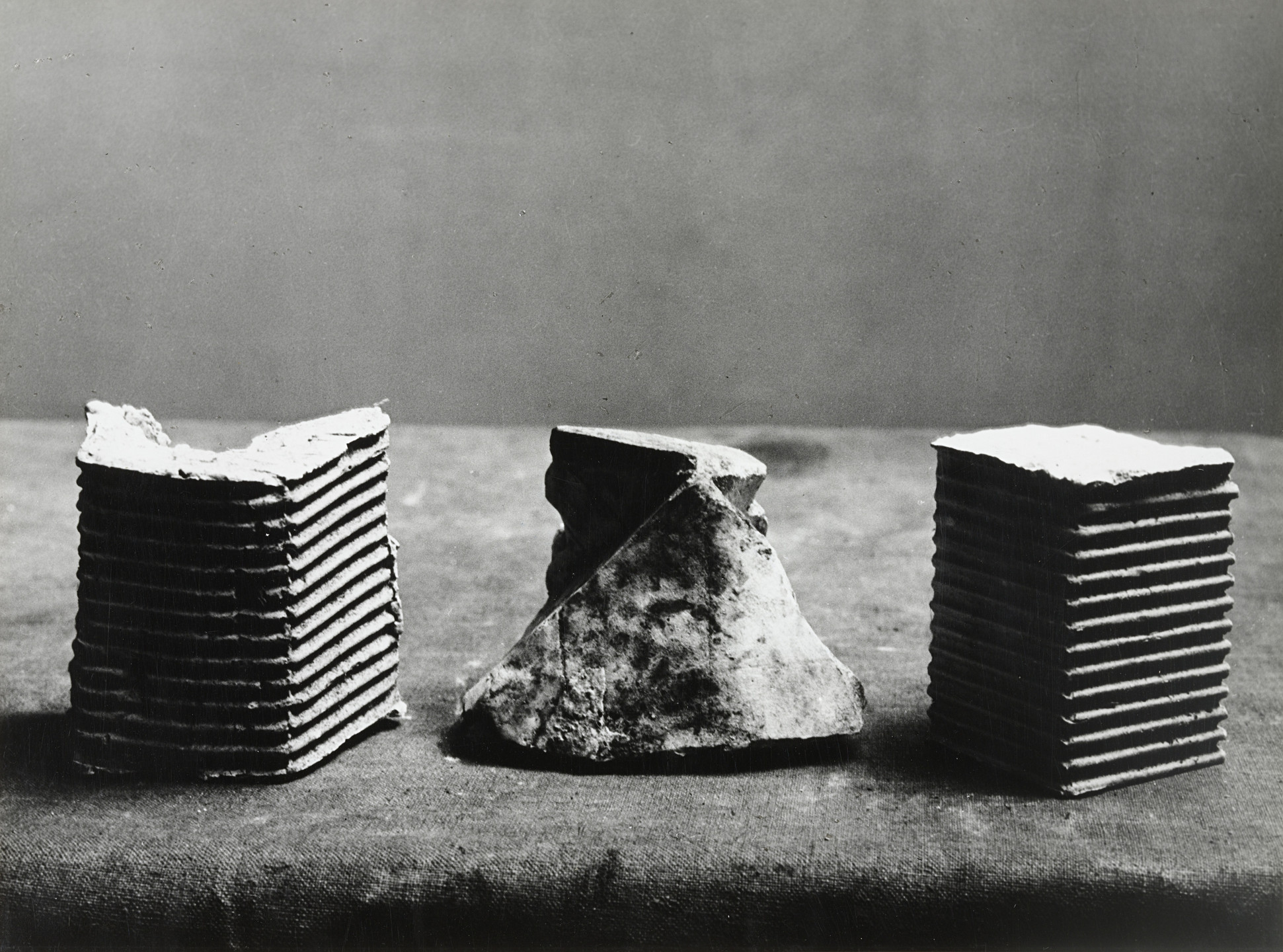 The image shows three sculptures on a gray surface with a gray background. The left and right sculptures are composed of stacked, ribbed slabs, while the middle sculpture is an irregularly shaped, rough piece. This is an AI generated description. To give feedback, email ai@moma.org.
