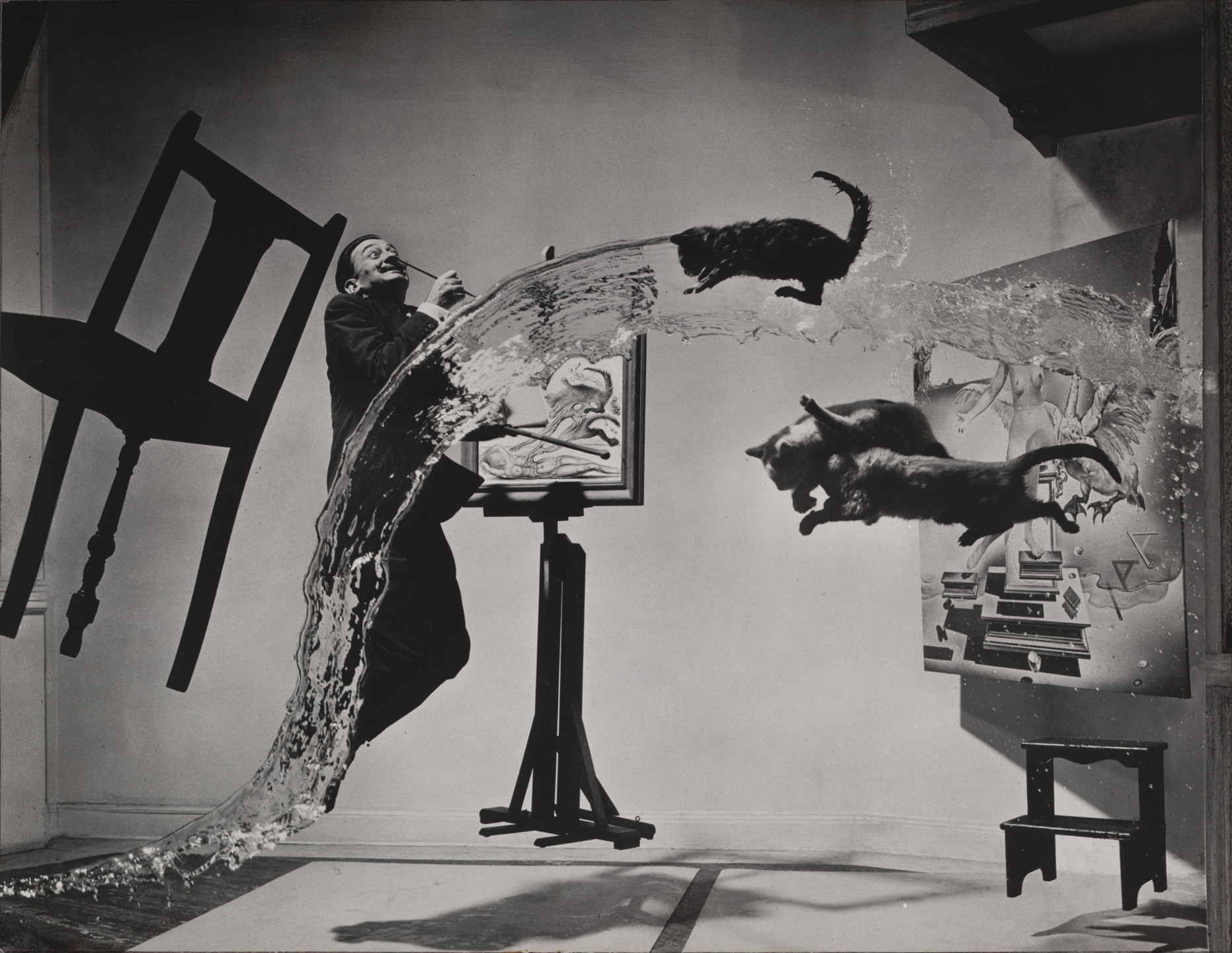 A person stands next to an easel, which holds a painting, while water and three cats are suspended mid-air. Surrounding them is a floating chair and a small table against a gray backdrop. This is an AI generated description. To give feedback, email ai@moma.org.