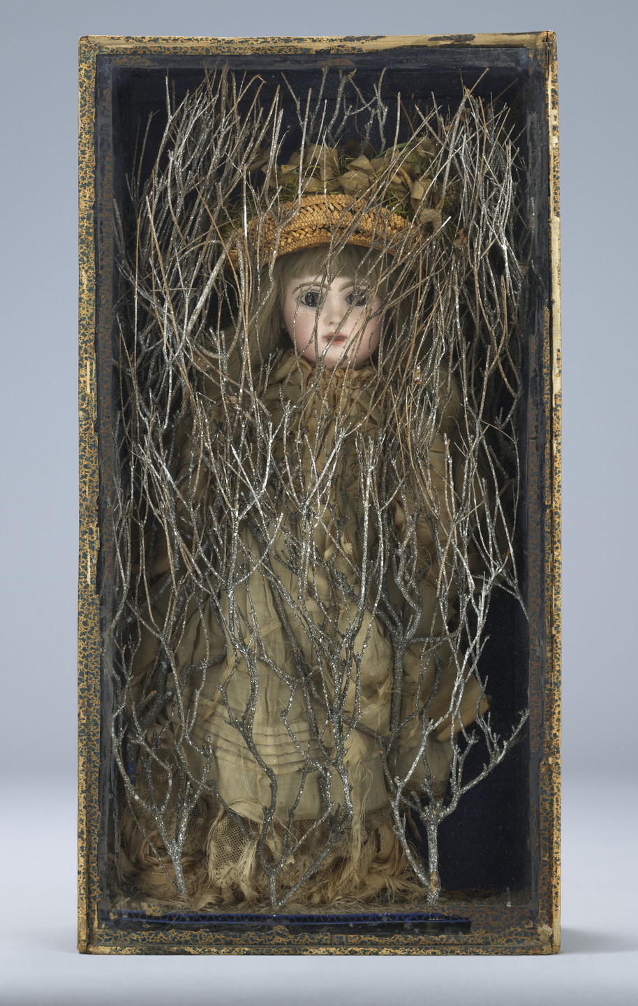 A doll with a porcelain face and blonde hair, dressed in brown and beige clothing, is encased in a box with intertwined silver branches obscuring its figure. The box has a worn, decorated edge. This is an AI generated description. To give feedback, email ai@moma.org.
