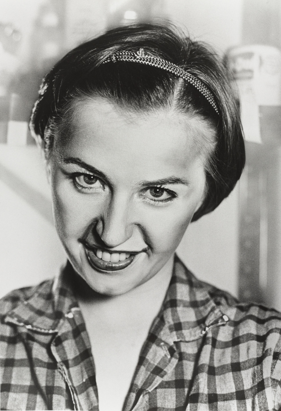 A black and white photo of a person with short hair wearing a headband and a checkered shirt. The person is smiling and looking directly at the camera. This is an AI generated description. To give feedback, email ai@moma.org.