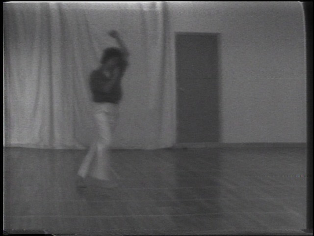 A person is blurred and in motion near the center, with an arm raised, standing on a shiny floor in a room. The backdrop features a curtain on the left and a closed door to the right. This is an AI generated description. To give feedback, email ai@moma.org.