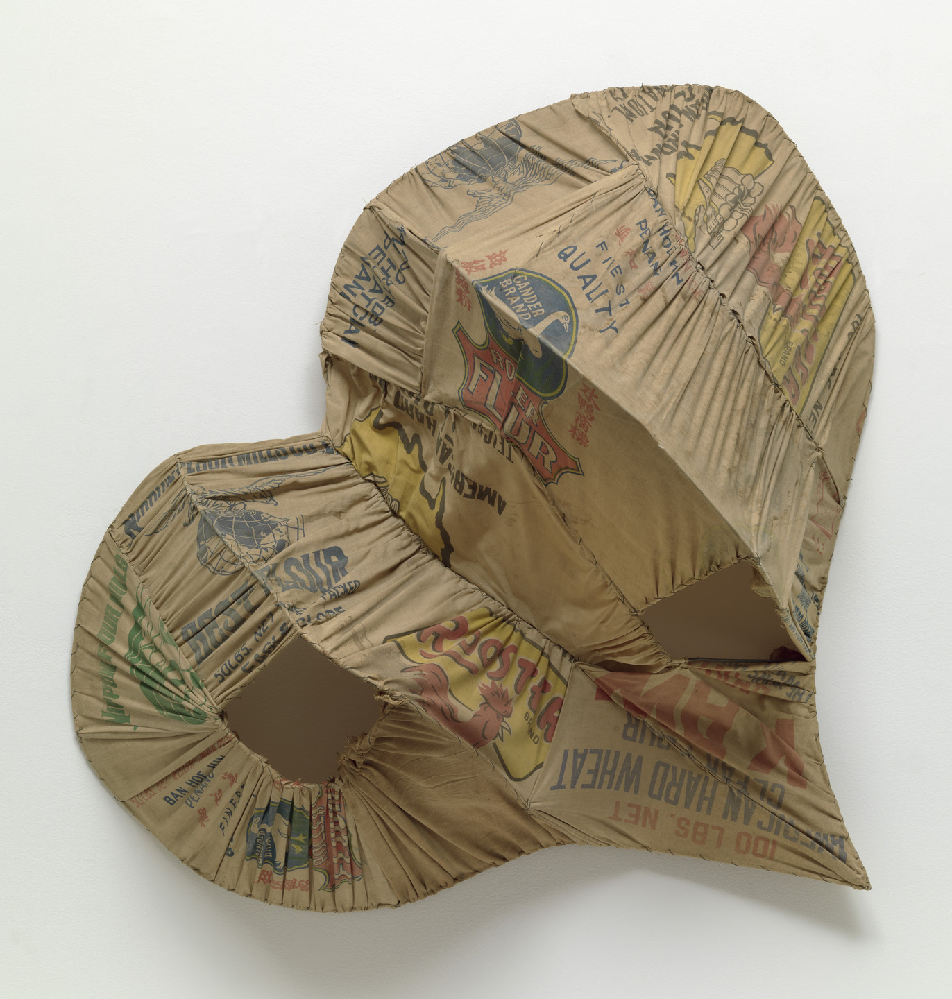 The image shows a large, irregularly shaped object made from sewn-together pieces of tan-colored fabric with various printed text and graphics in red, blue, yellow, and green. Some areas of the fabric are gathered and have small openings, creating a textured and layered appearance. This is an AI generated description. To give feedback, email ai@moma.org.
