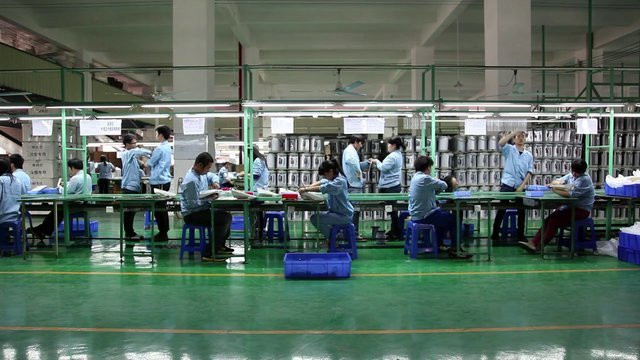 A group of people in light blue shirts works at a long table inside a spacious room with green floors and white walls. Shelves with organized items are in the background. This is an AI generated description. To give feedback, email ai@moma.org.