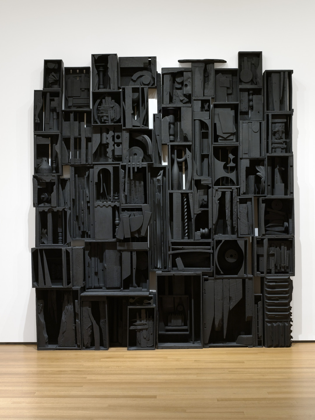 A wall-mounted assemblage composed of numerous black boxes filled with various objects and shapes. The piece is set against a white background with a light wood floor below. This is an AI generated description. To give feedback, email ai@moma.org.
