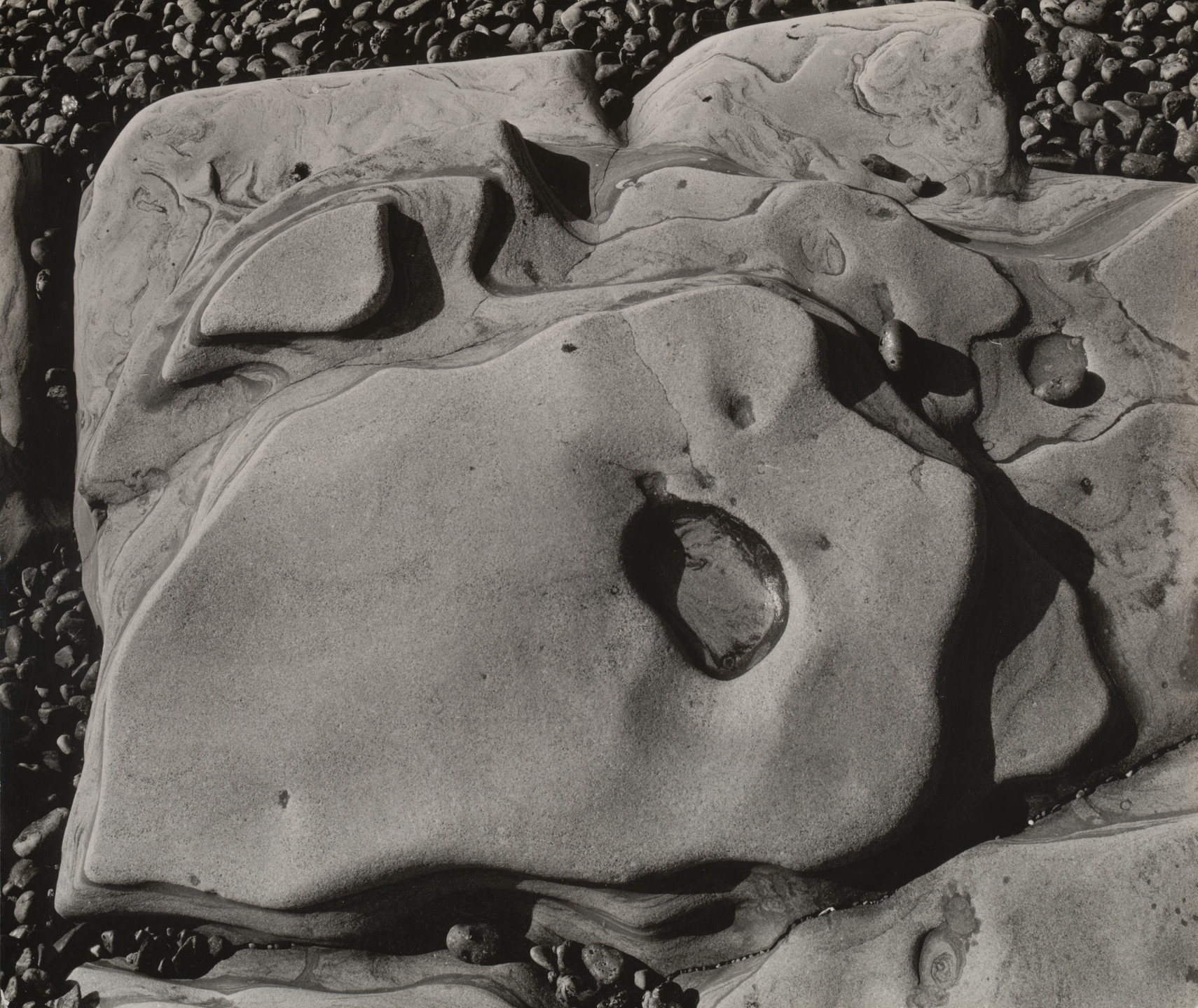 The image shows smooth, weathered rock formations with rounded shapes and deep grooves, predominantly in shades of grey. Smaller pebbles are scattered on the rocks and concentrated at the top edge. This is an AI generated description. To give feedback, email ai@moma.org.