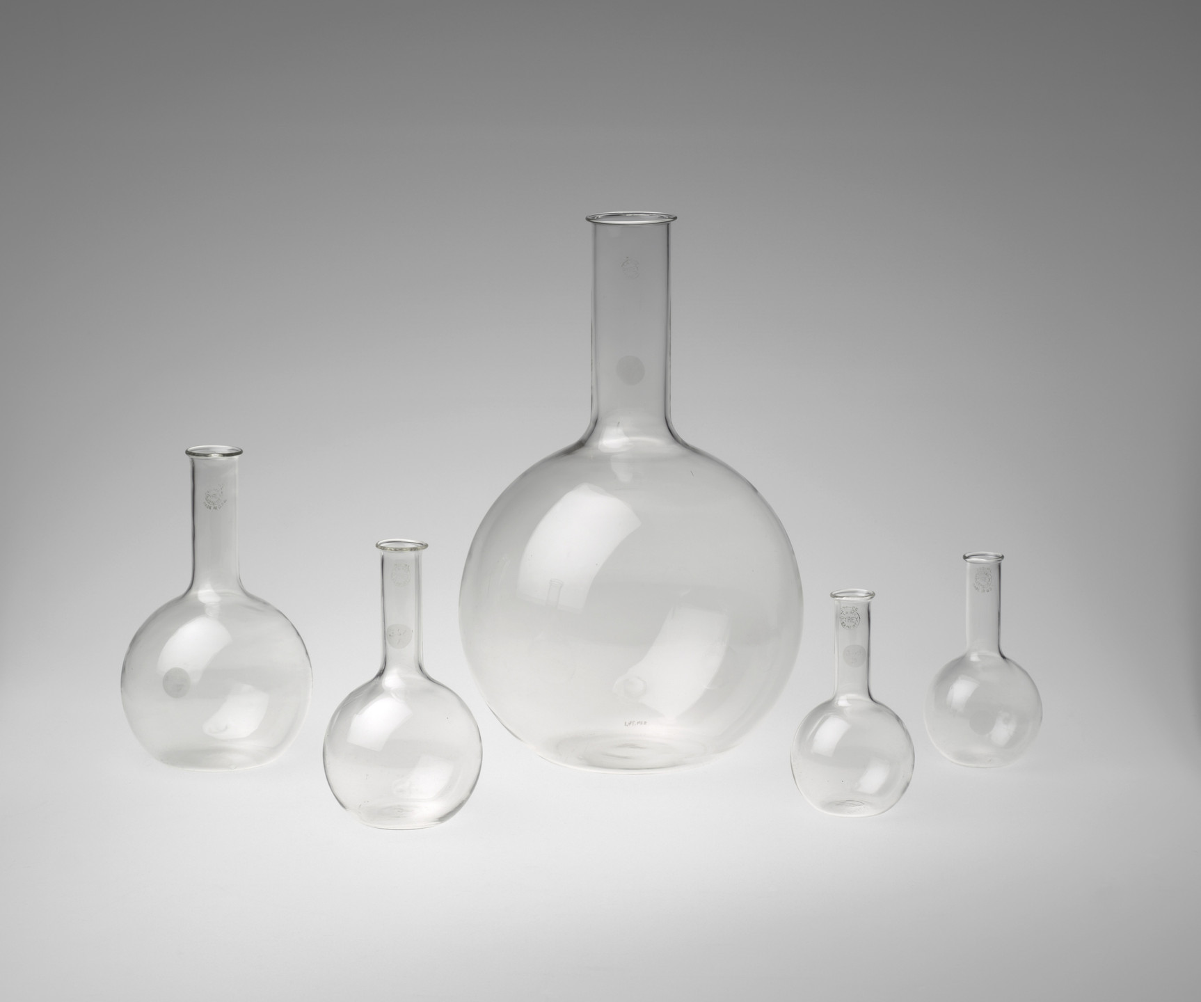 The image shows five clear glass flasks of various sizes with long necks arranged in a row against a gradient grey background. The largest flask is in the center, flanked by two medium-sized and two smaller ones on either side. This is an AI generated description. To give feedback, email ai@moma.org.