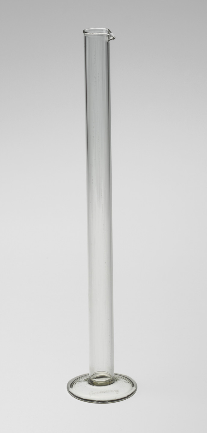 A tall, transparent glass cylinder with a flat, circular base is centered on a light gray background. The cylinder&#39;s top has a small spout for pouring. This is an AI generated description. To give feedback, email ai@moma.org.