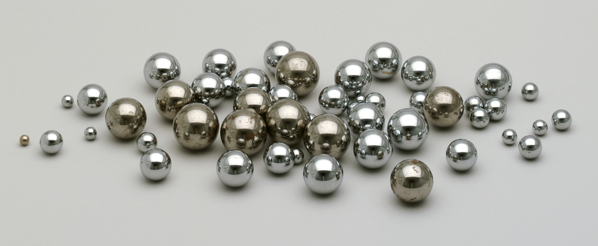 A scattered arrangement of silver and bronze ball bearings, varying in size, on a plain white background. The majority of the balls are shiny silver, with a few tarnished bronze ones. This is an AI generated description. To give feedback, email ai@moma.org.