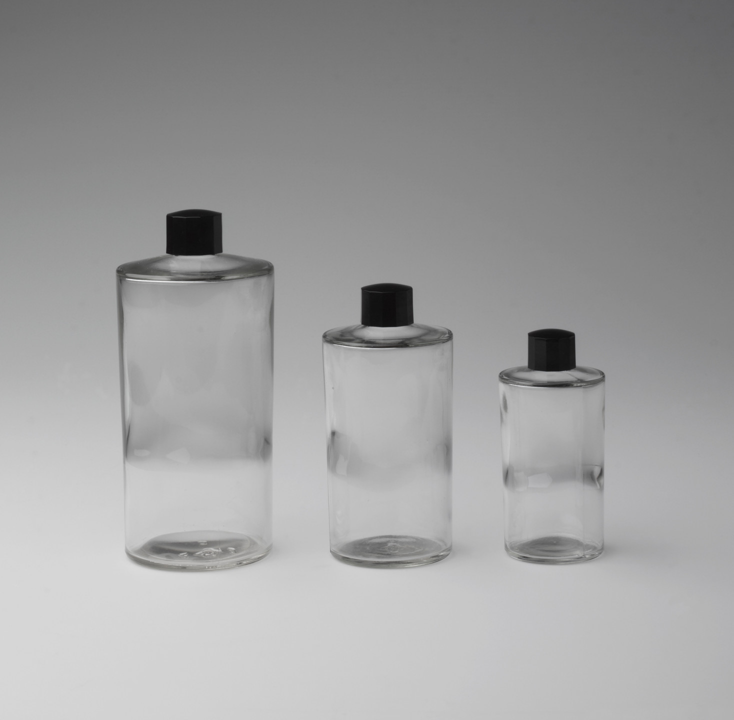 Three transparent cylindrical bottles of varying sizes with black caps are aligned in a row. The background gradient transitions from light gray at the bottom to a darker gray at the top. This is an AI generated description. To give feedback, email ai@moma.org.