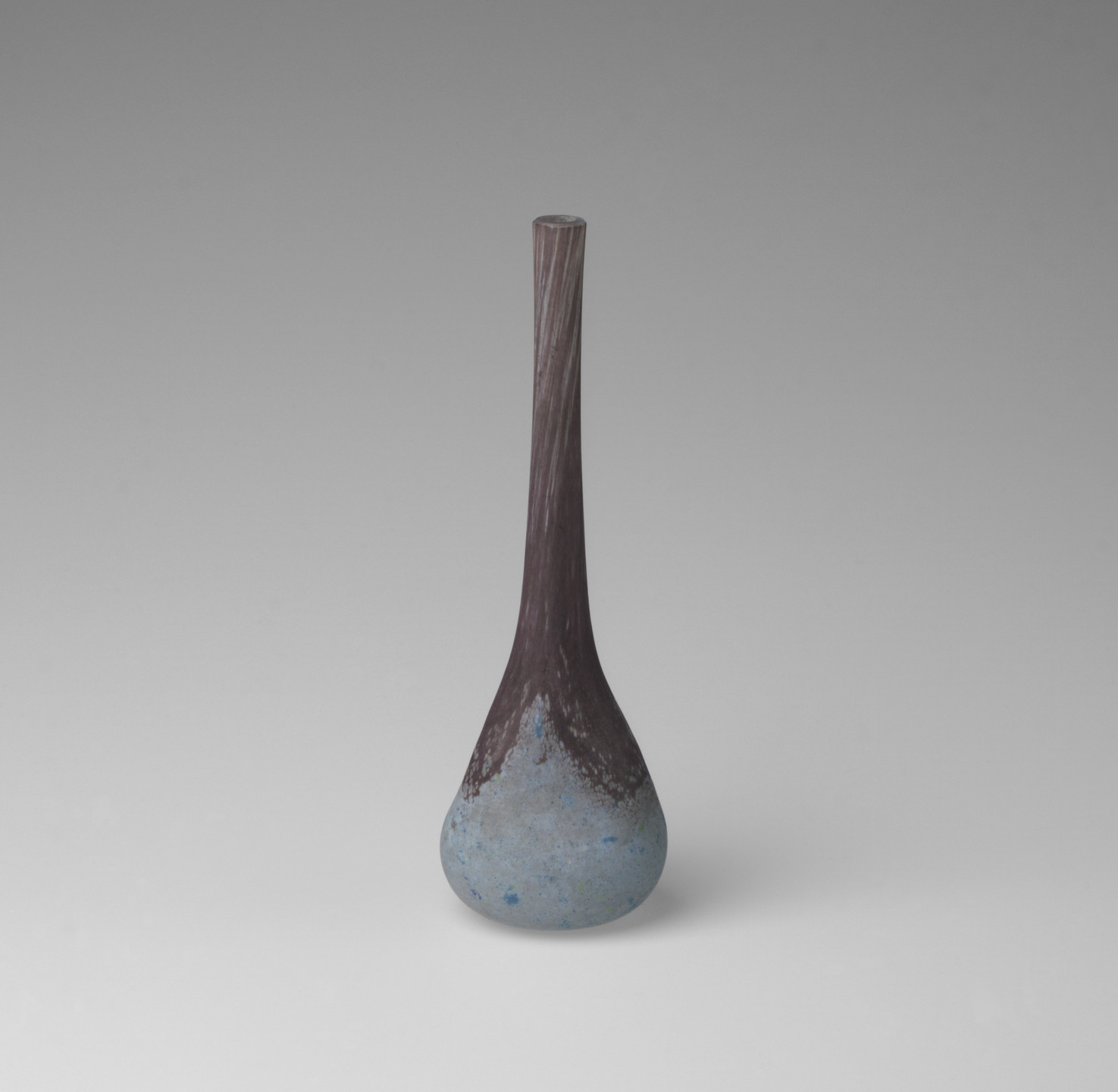 A tall, slender vase with a narrow neck and bulbous base blends brown at the top with speckled blue at the bottom, set against a light gray background. This is an AI generated description. To give feedback, email ai@moma.org.