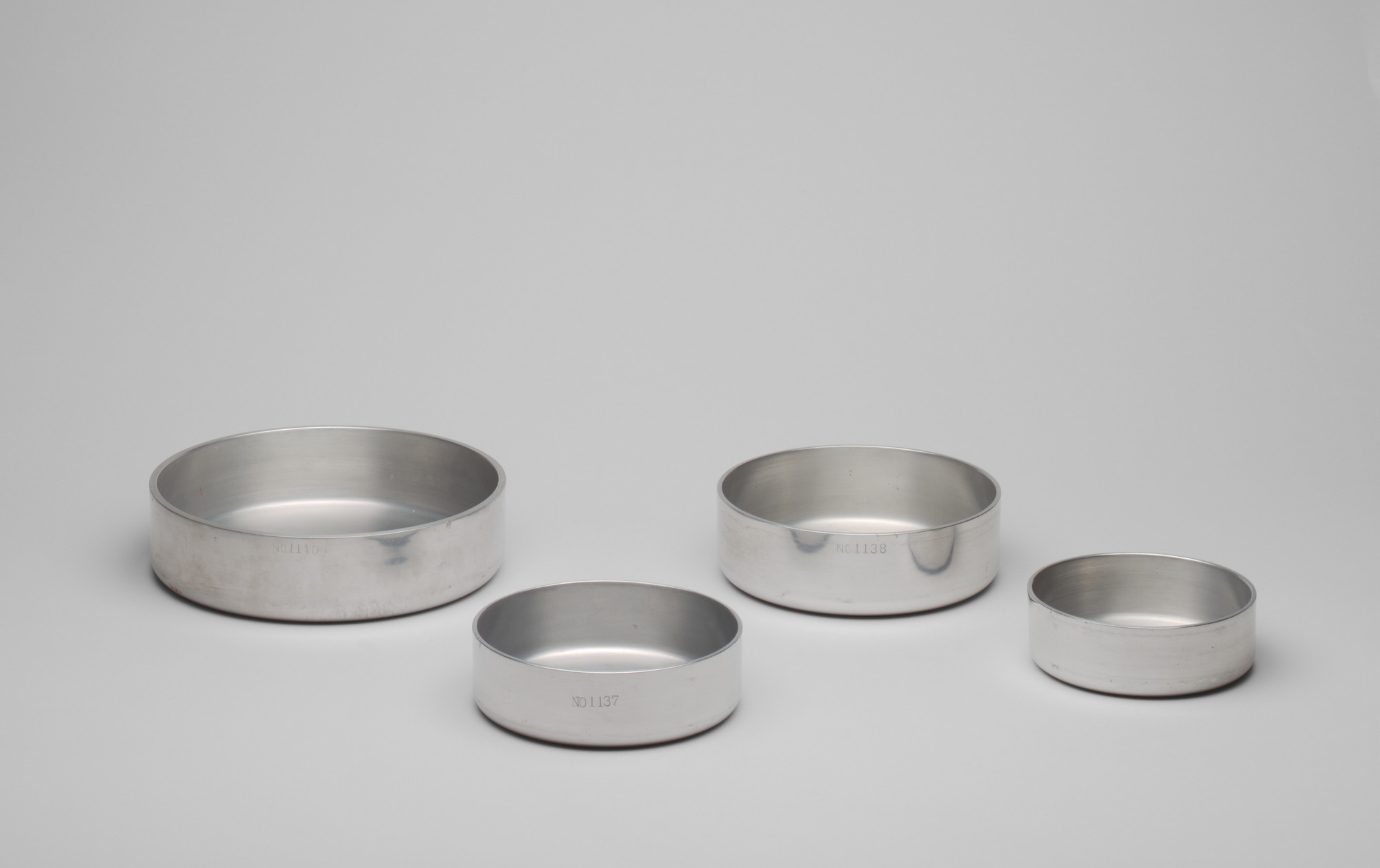 Four silver metal circular containers of different sizes are arranged in a line on a gray surface with a gray background. Each container has smooth walls and a flat bottom. This is an AI generated description. To give feedback, email ai@moma.org.