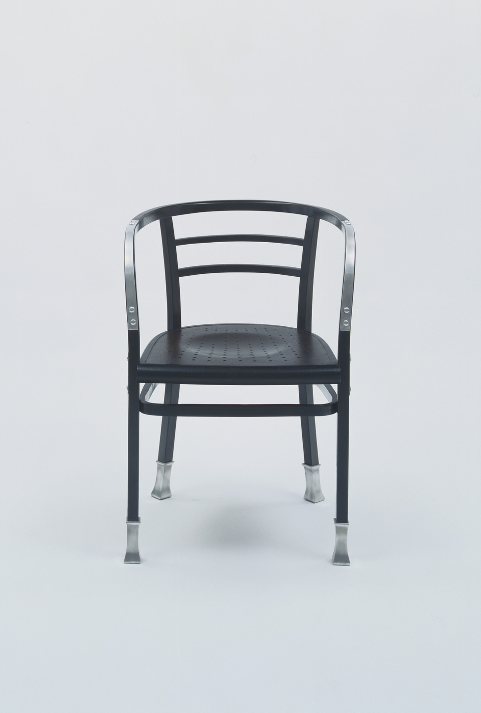 A black chair with a perforated seat, curved backrest, and thin legs ending in metal caps is centered against a plain white background. Silver accents are visible on the backrest and legs. This is an AI generated description. To give feedback, email ai@moma.org.