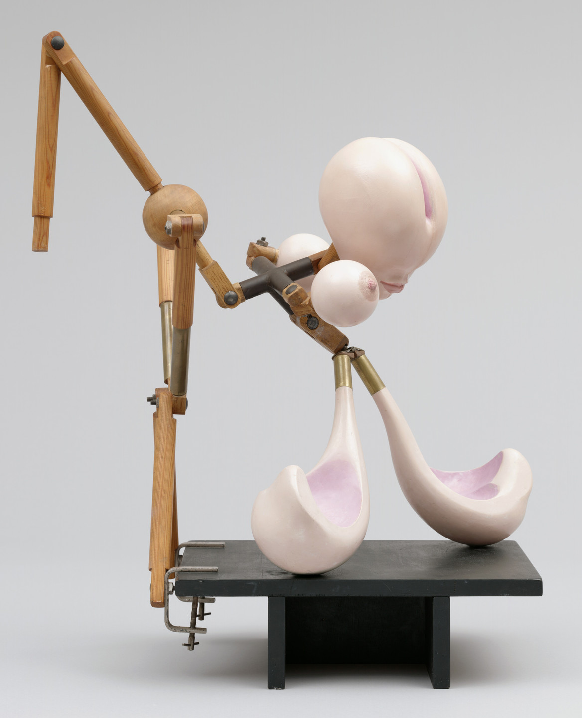 A sculpture with pale pink bulbous forms and wooden, jointed mechanical limbs is mounted on a black rectangular base. The limbs have metallic and brass connections, giving a structured mechanical appearance. This is an AI generated description. To give feedback, email ai@moma.org.