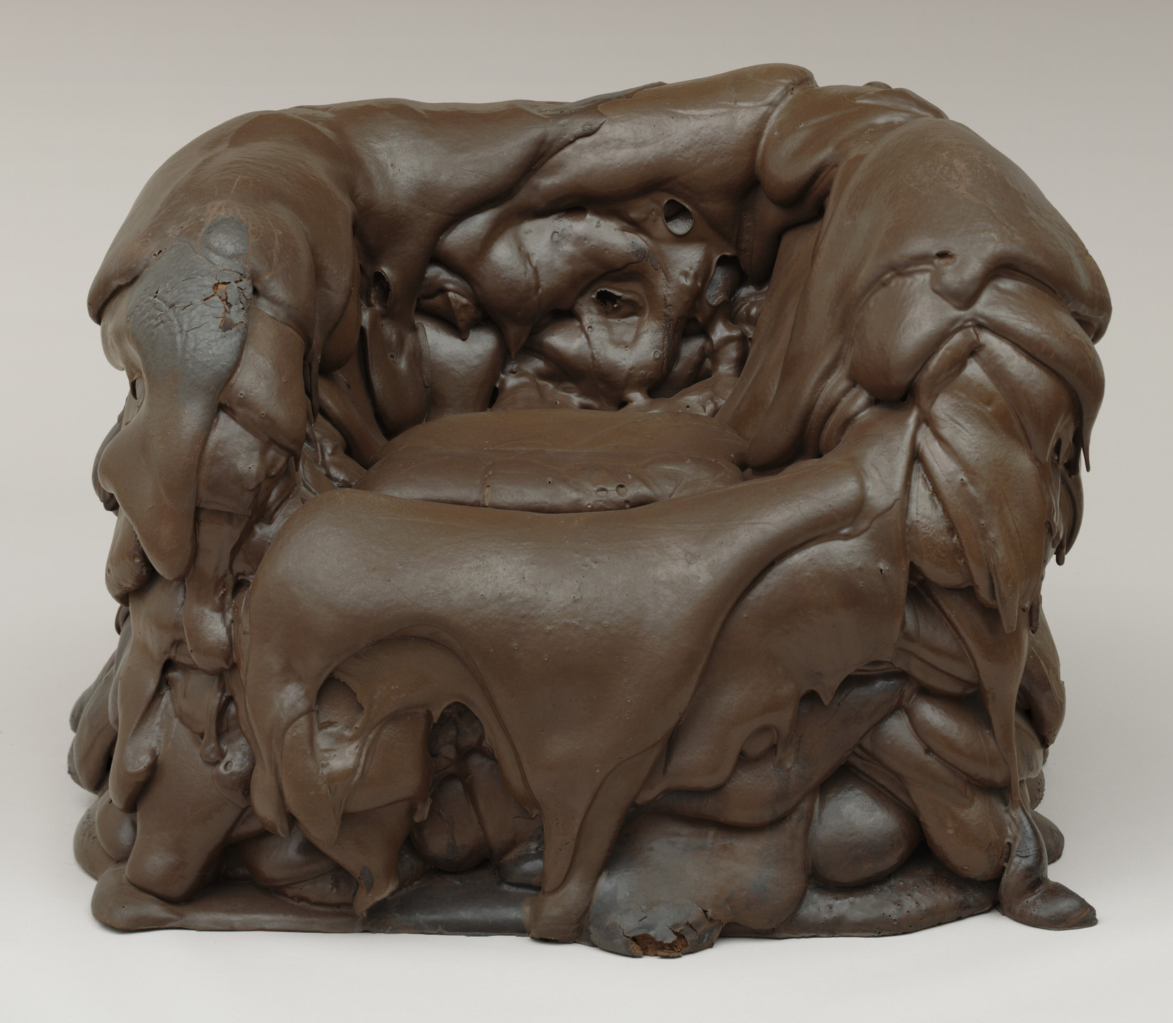 A brown chair appears to be made of a soft, melting material with uneven, flowing textures and folds like melting chocolate or flowing lava. This is an AI generated description. To give feedback, email ai@moma.org.