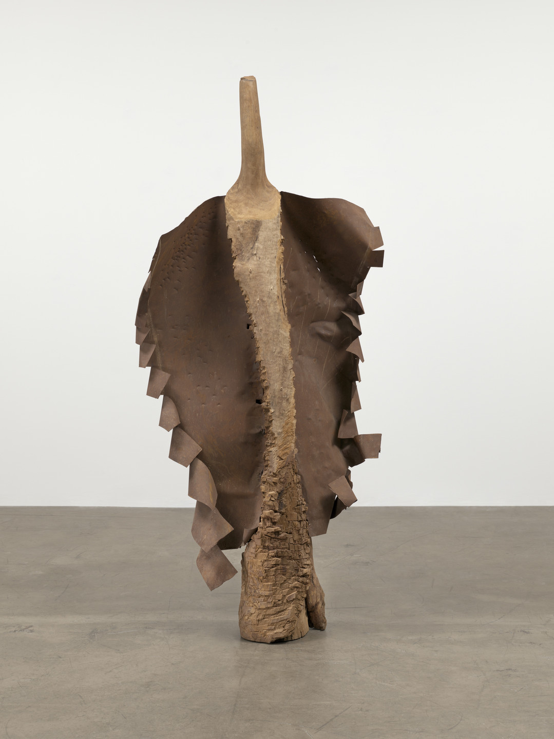 A sculpture made of wood and metal in various shades of brown stands on a gray floor. A wide sheet of brown metal is attached to the wood, its edges are cut into jagged strips. This is an AI generated description. To give feedback, email ai@moma.org.