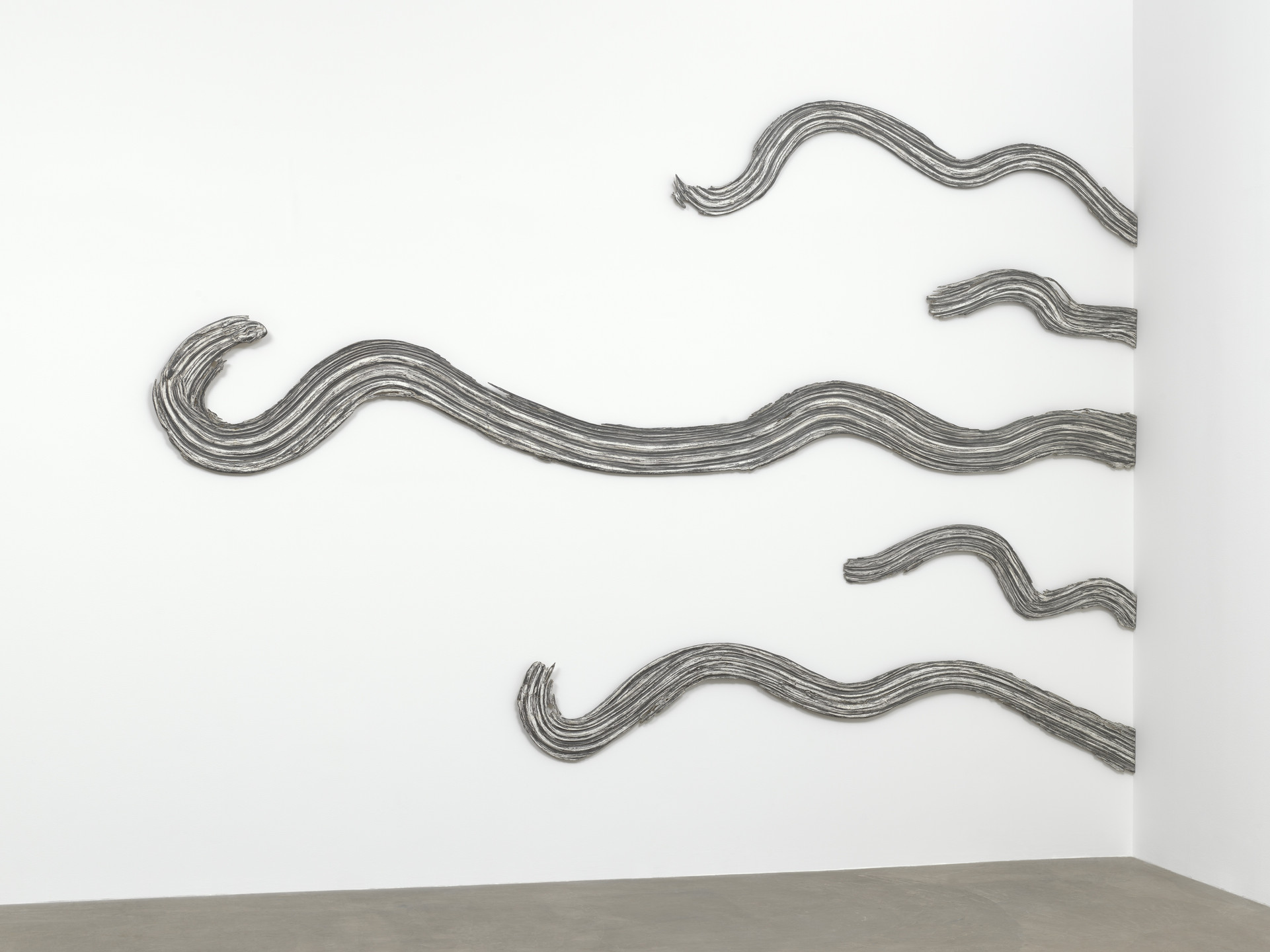Gray, wavy lines are spread horizontally across a white wall, curving and overlapping at some points. The wall and adjoining floor are otherwise bare, creating a minimalist look. This is an AI generated description. To give feedback, email ai@moma.org.