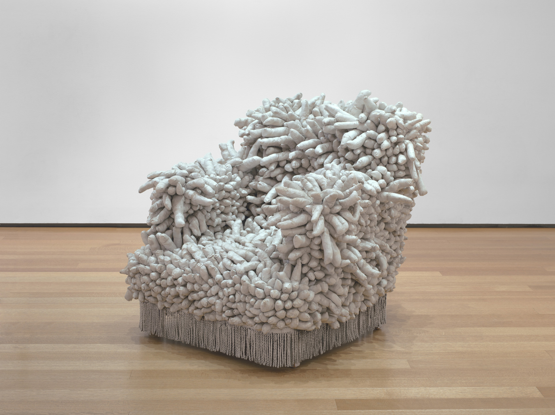 A chair covered in numerous soft, finger-like white protrusions stands on a wooden floor with a plain white wall backdrop. It has a fringe along the bottom edge. This is an AI generated description. To give feedback, email ai@moma.org.