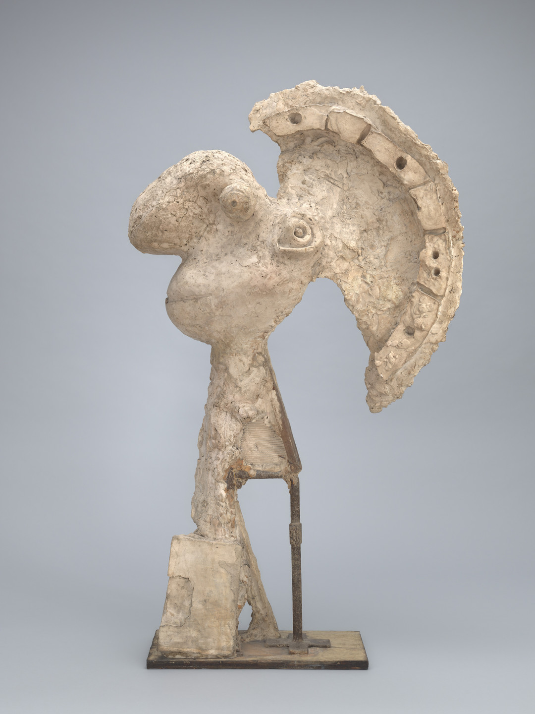 A beige, textured sculpture with a prominent, curved feature at the top, resembling a headdress, stands on a base. The background is a plain light gray. This is an AI generated description. To give feedback, email ai@moma.org.
