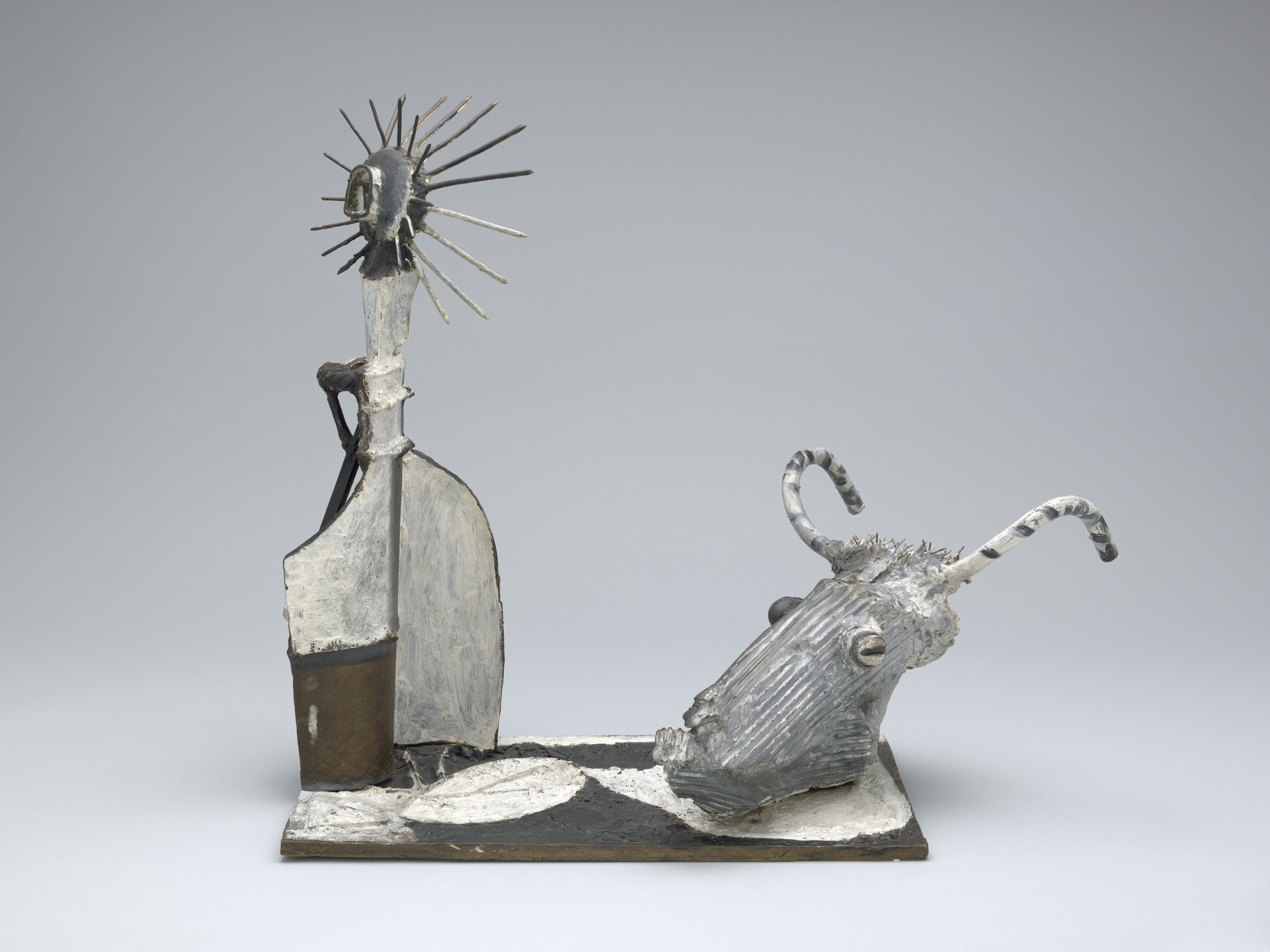 A sculpture features a spike-headed figure on the left, attached to a flat base, with a ribbed, animal-like head with curved horns on the right. The predominant colors are gray, white, and brown. This is an AI generated description. To give feedback, email ai@moma.org.