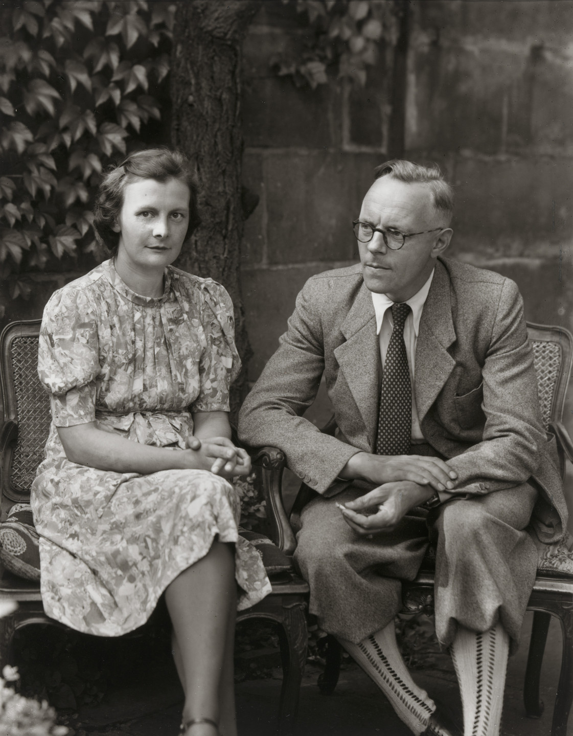 Two individuals sit side by side on wooden chairs in a garden; one wears a floral dress, and the other a suit with patterned socks. The background includes ivy and a stone wall. This is an AI generated description. To give feedback, email ai@moma.org.