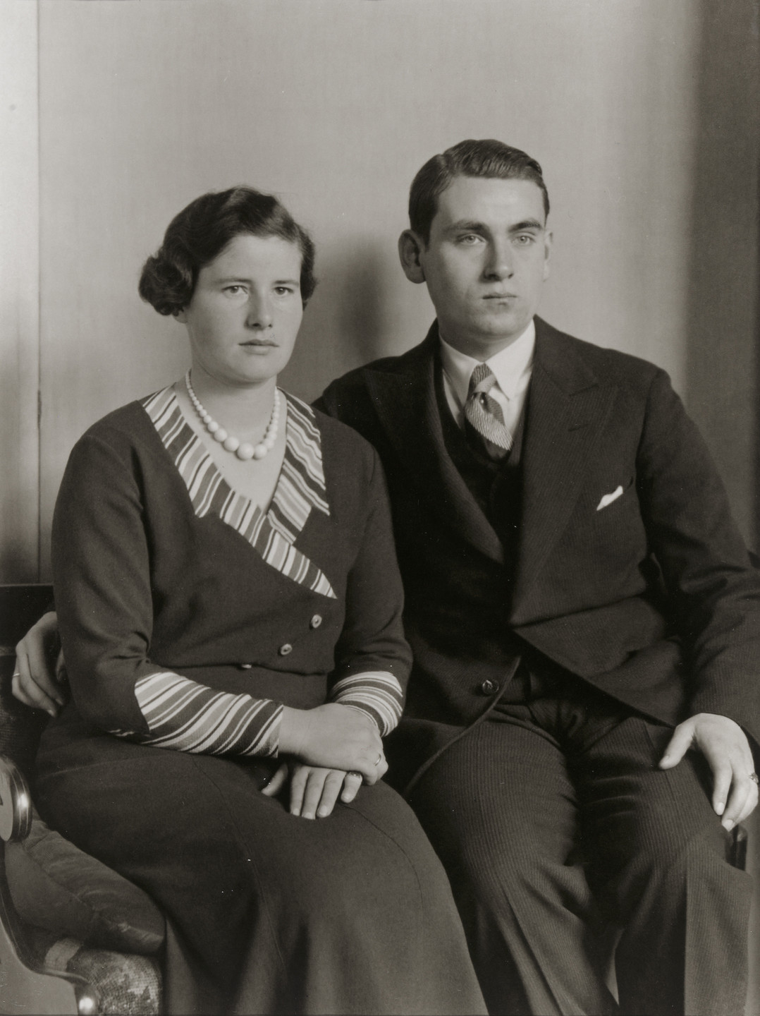 A black and white photo of two people seated side-by-side, with one wearing a dark suit and the other in a dark dress with striped accents and a pearl necklace. Both have serious expressions and are holding hands. This is an AI generated description. To give feedback, email ai@moma.org.