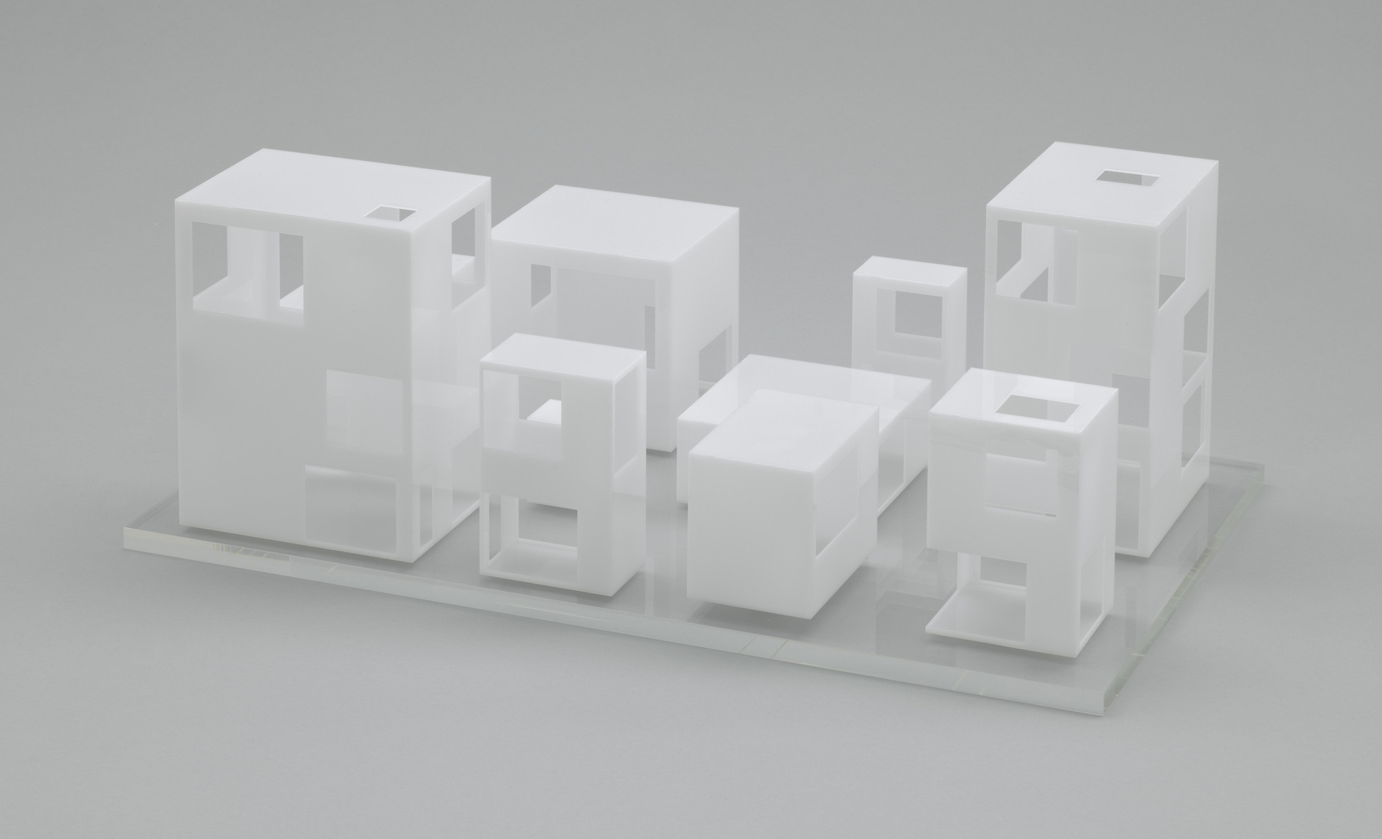 The image shows a model of white, geometric, box-like structures of various sizes on a flat, reflective surface. Each structure has cut-out square windows and rectangular openings. This is an AI generated description. To give feedback, email ai@moma.org.