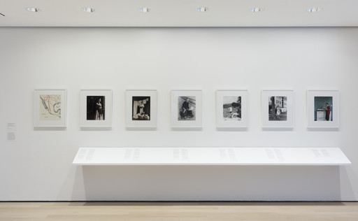 A white wall displays seven framed works, six black-and-white images and one color photograph. Below the frames is a long, white shelf with documents laid out for viewing. This is an AI generated description. To give feedback, email ai@moma.org.